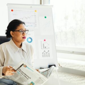 Confident businesswoman reading newspaper and analyzing market trends in a modern office setting.